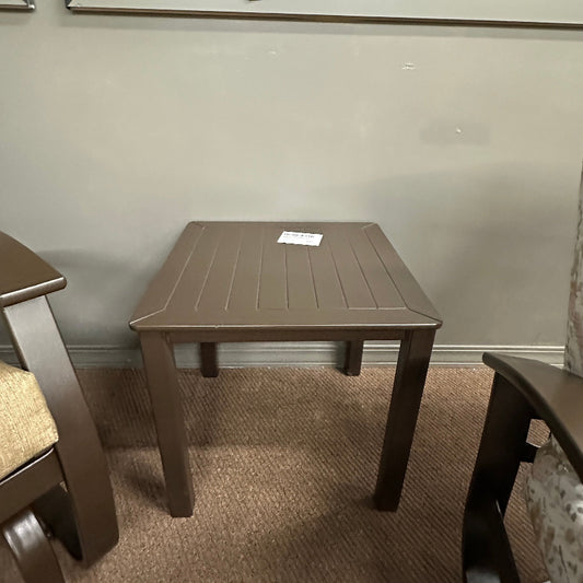 A square brown plastic side table with a slatted top and four legs, shown in a furniture store setting on a carpeted floor against a gray slatwall background.