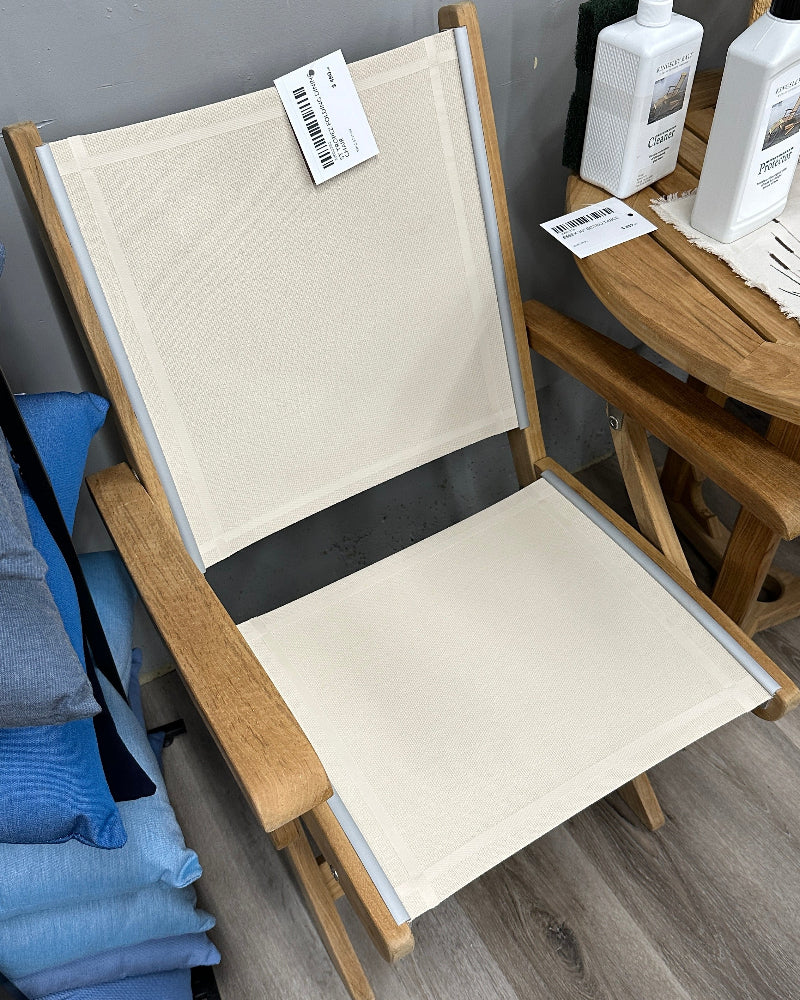 A wooden folding beach chair with cream canvas fabric, shown in a store setting on a gray wood floor next to a table with bottles.