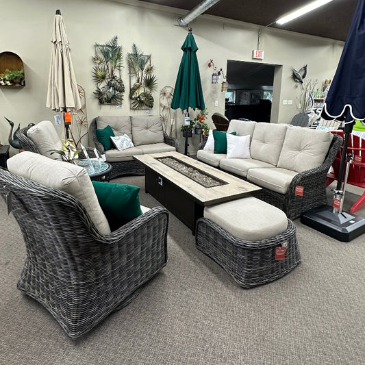 Outdoor patio furniture set in a showroom, featuring a gray wicker sectional sofa, matching armchair, ottoman, and a rectangular fire pit coffee table with a light stone top.
