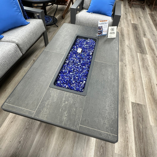 Gray fire pit table with blue decorative stones on a wooden floor, surrounded by furniture.