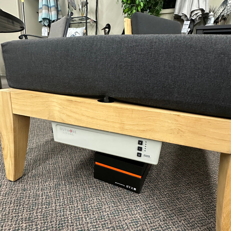 A close-up of a modern outdoor sofa or chaise lounge with a dark gray fabric cushion on a light oak wood frame, shown in a showroom setting with a patterned carpet and patio umbrellas in the background.