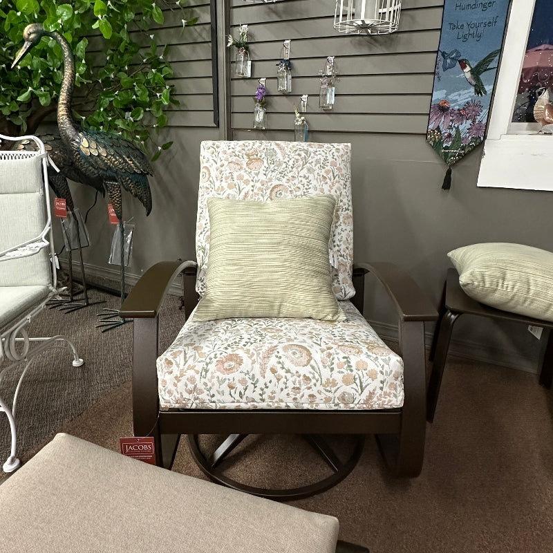 A dark wood outdoor swivel armchair with a cream and pink floral patterned fabric and a light green pillow, shown in a store display with an ottoman in the foreground.