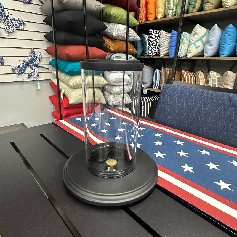 A black cylindrical table lamp with a clear glass shade and a circular black base, displayed on a dark slatted table in a furniture showroom. An American flag-patterned runner is visible underneath, with shelves of colorful pillows and cushions in the background.