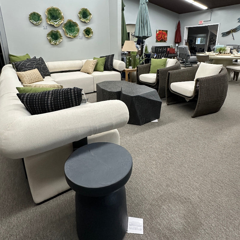 A modern outdoor lounge set displayed in a showroom, featuring a white curved sectional sofa with green and black pillows, two dark gray armchairs, black coffee tables, and a round black side table on a gray carpeted floor.