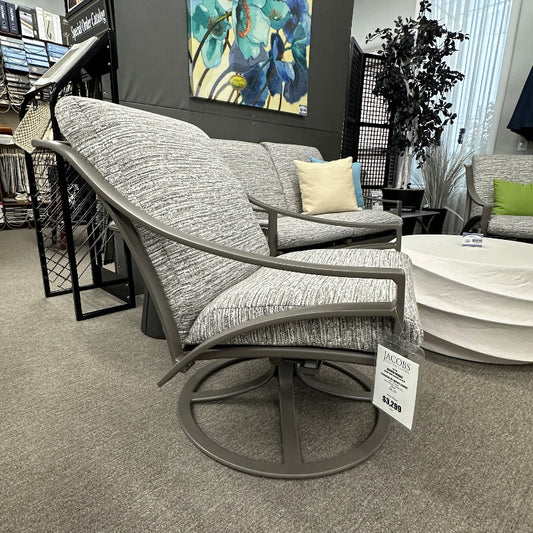 Gray swivel chair in a showroom setting with a painting on the wall and a couch in the background.