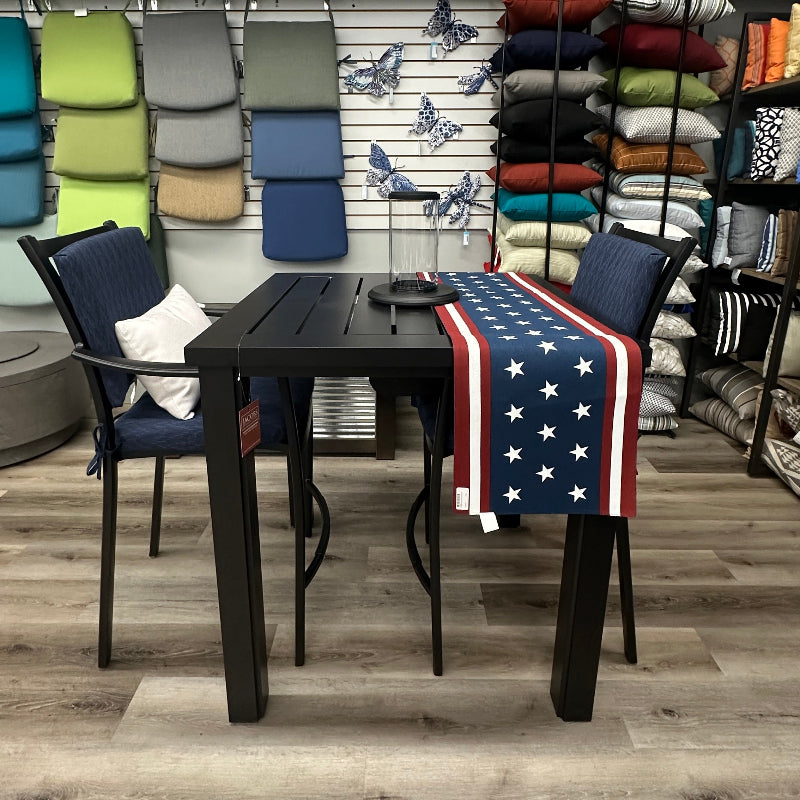 A dark outdoor dining table with matching armchairs in a showroom, featuring a red, white, and blue American flag patterned throw blanket draped over the front right chair. Shelves in the background are filled with colorful pillows and folded home textiles.
