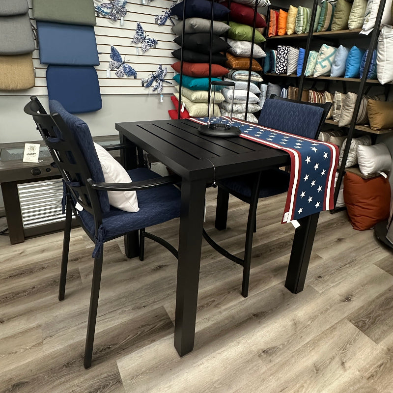 A dark wood outdoor dining table with two navy blue armchairs in a furniture showroom. Shelves in the background are filled with colorful pillows and folded home textiles.