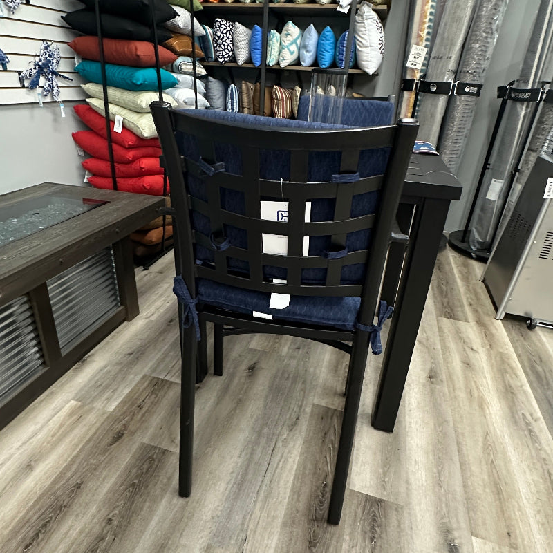 A dark wood dining chair with a blue woven seat cushion and back strap, positioned in a furniture showroom with wood floors and shelves of pillows and textiles in the background.