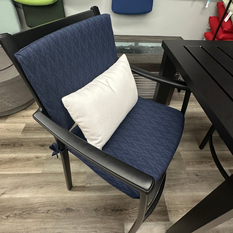 A navy blue outdoor patio armchair with a white accent pillow, shown next to a black slat-top table on a wood-look floor. A fabric swatch display is visible on the wall behind.
