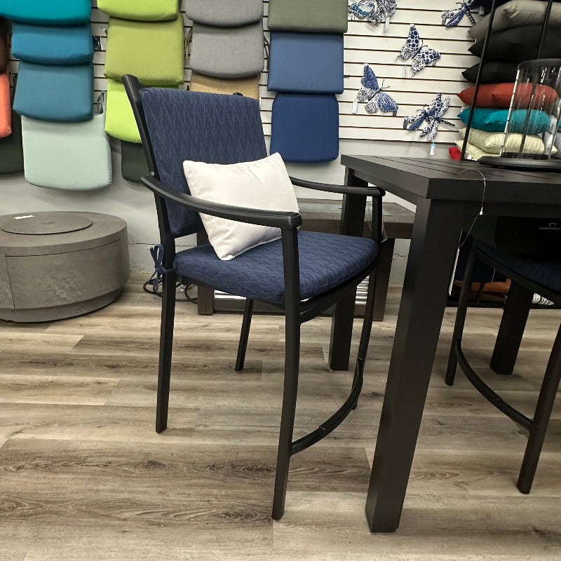 A navy blue outdoor patio dining chair with a white accent pillow, set next to a dark dining table on a light wood floor in a furniture showroom.