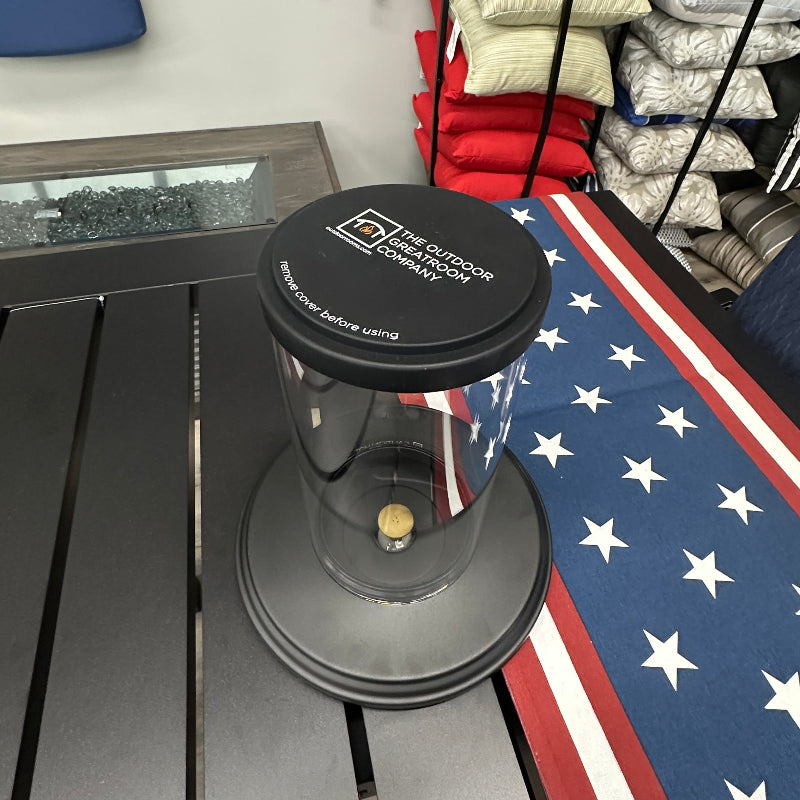 A clear glass patio umbrella canopy with a black top cap and base, branded 'The Patio Company', displayed on a gray slatted table in a retail showroom next to an American flag runner.