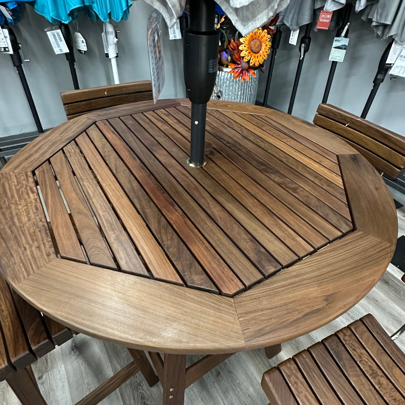 A round outdoor patio dining set with a brown wooden slatted table, matching chairs, and a gray striped umbrella in a store display.