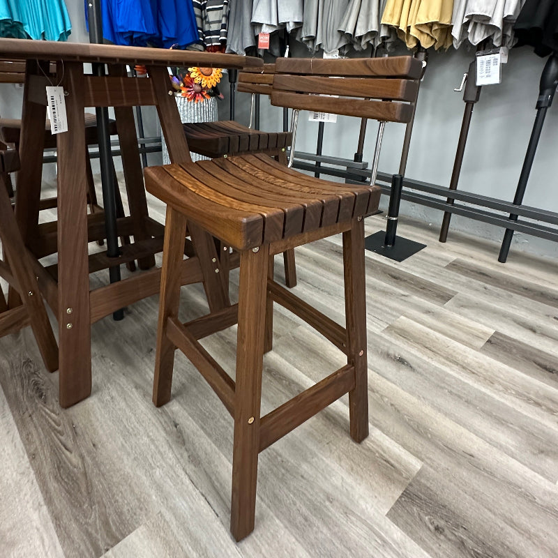 A wooden outdoor bar stool with a slatted seat and back, shown next to a matching wooden table in a retail store setting with gray wood-look flooring and patio umbrellas in the background.