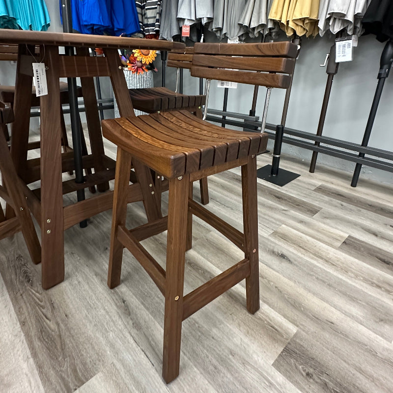 A wooden outdoor bar stool with a slatted seat and back, shown next to a matching wooden table in a retail store setting with gray wood-look flooring and patio umbrellas in the background.