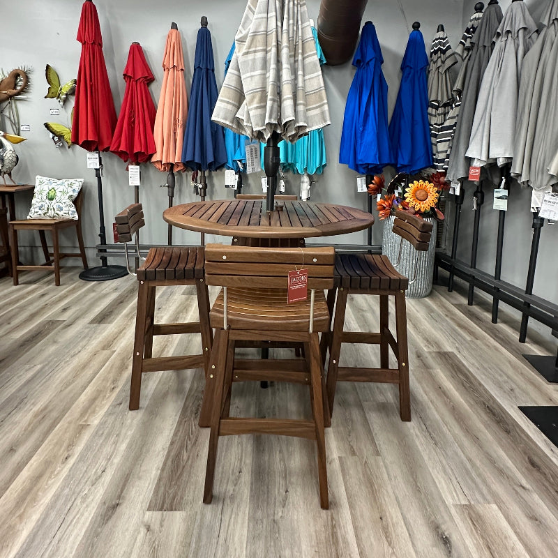 A wooden outdoor dining set consisting of a round slatted table and four matching bar stools, displayed in a furniture showroom with other patio umbrellas and decor in the background.