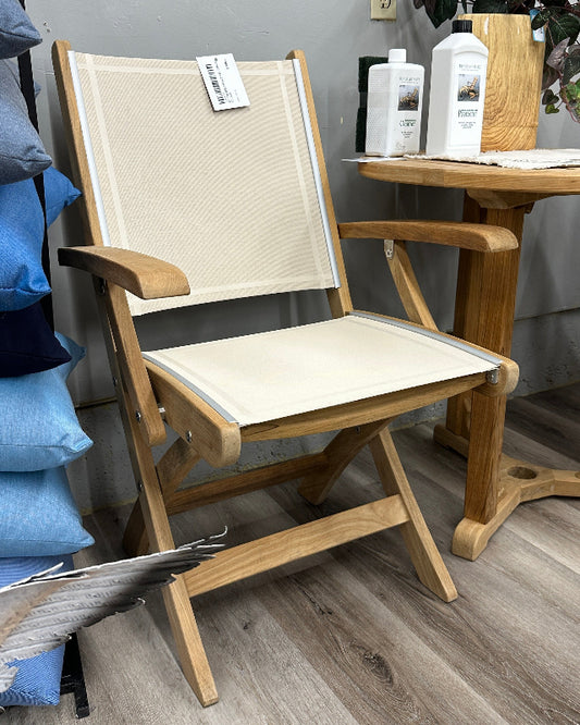 A folding outdoor-style chair with a light beige canvas seat and back, supported by a natural wood frame, shown in a home decor store setting next to a wooden side table.