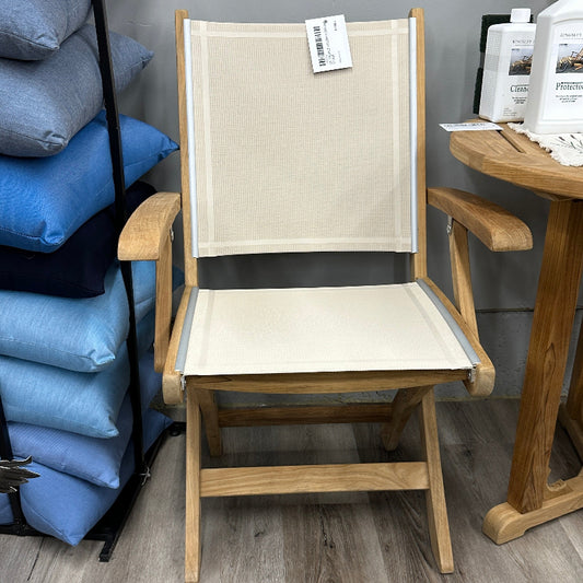 A wooden folding chair with a beige canvas seat and backrest, shown in a furniture store setting with a gray wall and wood-look floor.