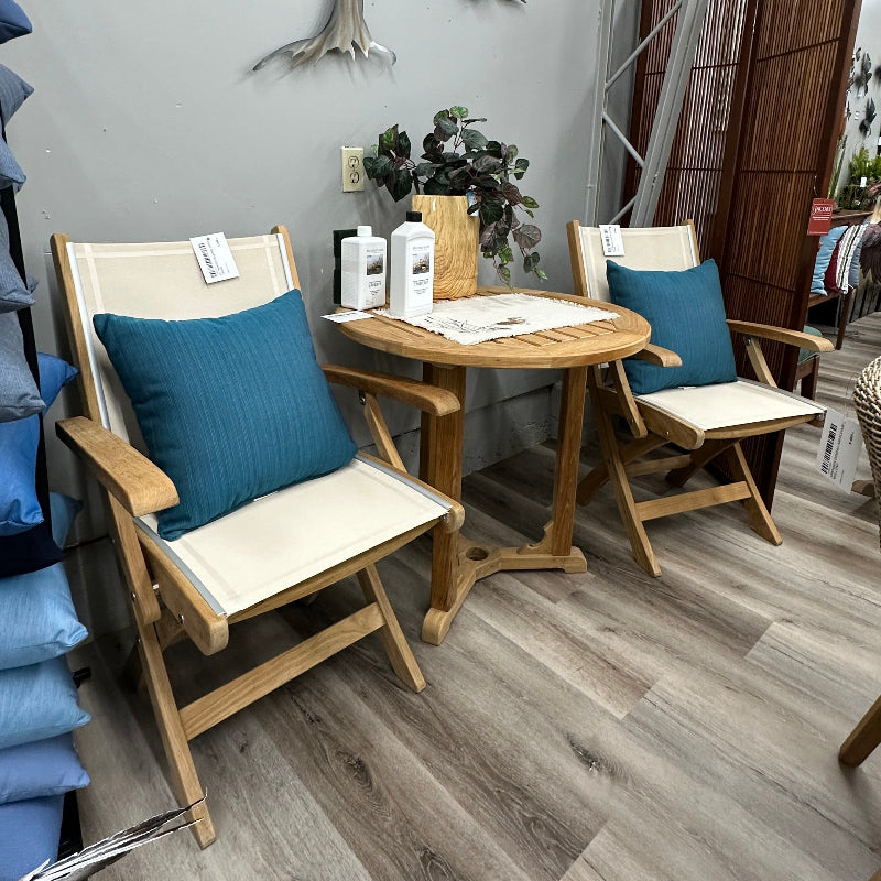 A styled interior scene showing two wooden folding chairs with cream fabric seats and blue pillows around a small round wooden table, set on a wood-look floor against a gray wall with a decorative tree sculpture.