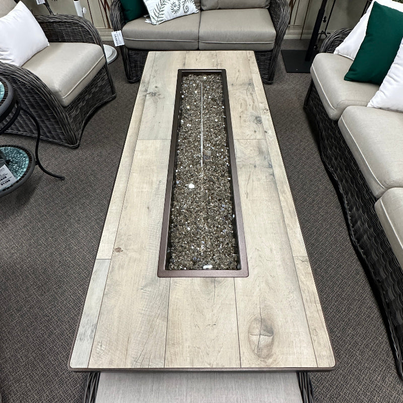 A rectangular outdoor fire pit table with a light wood-look top and a dark metal center channel filled with dark gravel, shown in a patio furniture showroom.