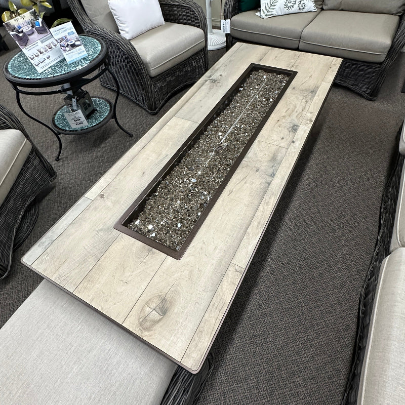 A rectangular outdoor fire pit coffee table with a light wood-look top and a dark metal firebox insert, shown in a furniture showroom with patio seating around it.