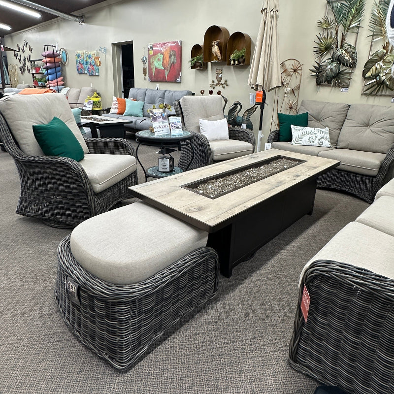 Outdoor wicker patio furniture set in a showroom, including a rectangular fire pit table with a light wood top, two gray wicker armchairs with off-white cushions, and a matching ottoman, surrounded by a gray sectional sofa.