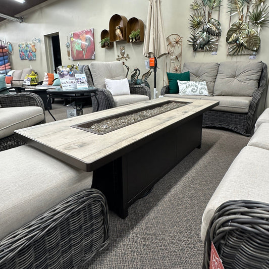 A modern outdoor patio conversation set featuring a rectangular fire pit coffee table with a light wood top and black base, surrounded by gray wicker furniture with light beige cushions in a showroom setting.