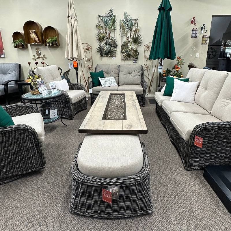Outdoor wicker patio furniture set in a showroom, including a sofa, armchair, ottoman, coffee table, and matching chairs with cream cushions and green accents.