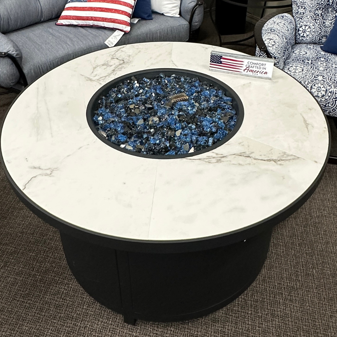Round outdoor fire pit table with a white marble-patterned top and black base, filled with blue and black fire stones. The table is shown in a showroom setting with a gray tufted sofa and patterned armchair in the background.