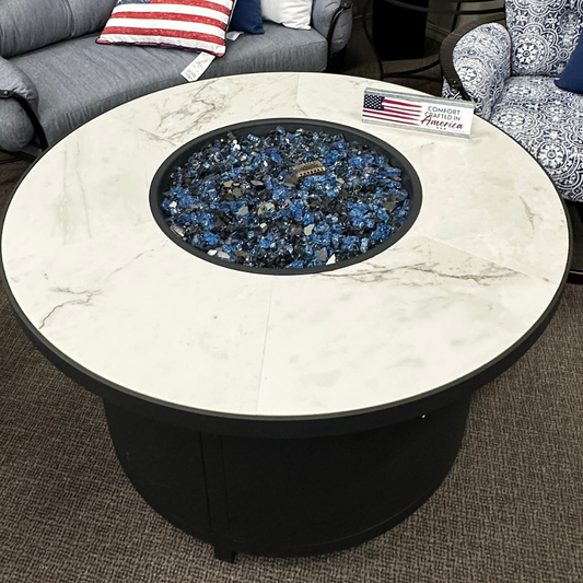 Round outdoor fire pit table with a white marble-patterned top and black base, filled with blue and black fire stones. The table is shown in a showroom setting with a gray tufted sofa and patterned armchair in the background.