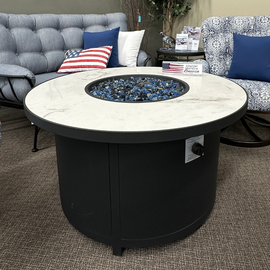 A round outdoor fire pit table with a white marble-patterned top and a black cylindrical base filled with blue decorative stones, shown in a furniture showroom between two sofas.