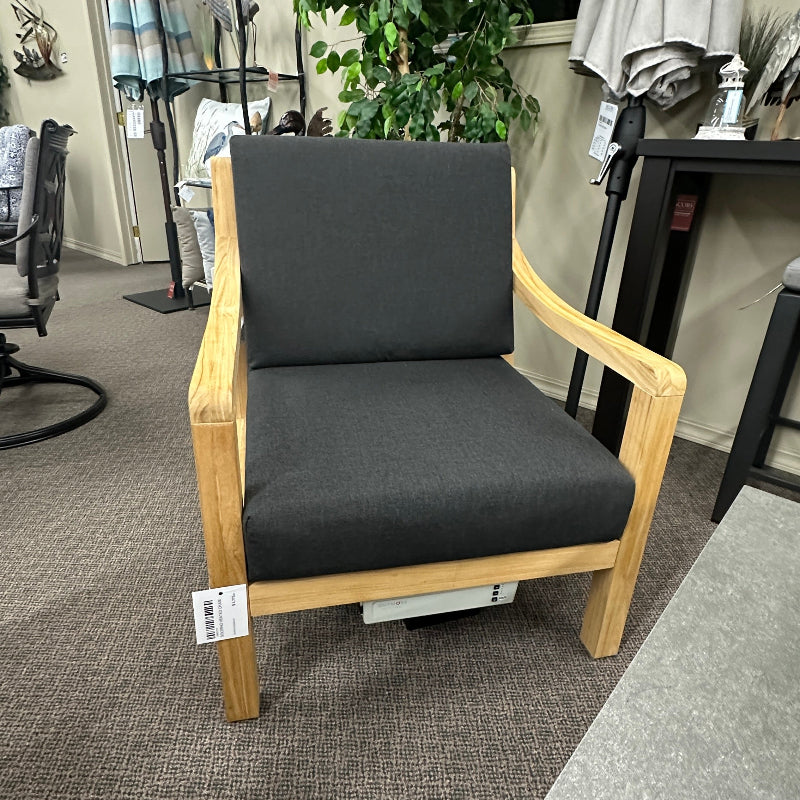 A wooden armchair with a dark gray upholstered seat and back cushion, photographed in a furniture showroom or office setting with carpet, plants, and other furniture visible.