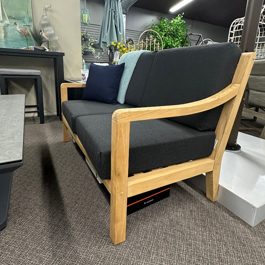 A modern wooden loveseat with dark gray fabric cushions and two accent pillows, set in a furniture showroom on a textured gray carpet.