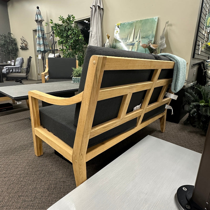 A wooden outdoor sofa with a light natural wood frame and dark gray cushions, shown in a furniture showroom setting.
