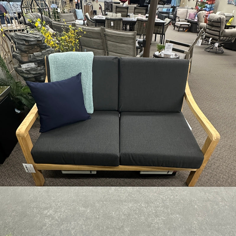 A two-seat outdoor loveseat with dark gray cushions and a light wood frame, shown in a furniture store. The loveseat includes a navy blue throw pillow and a light blue accent pillow, with a patio umbrella and other furniture visible in the background.