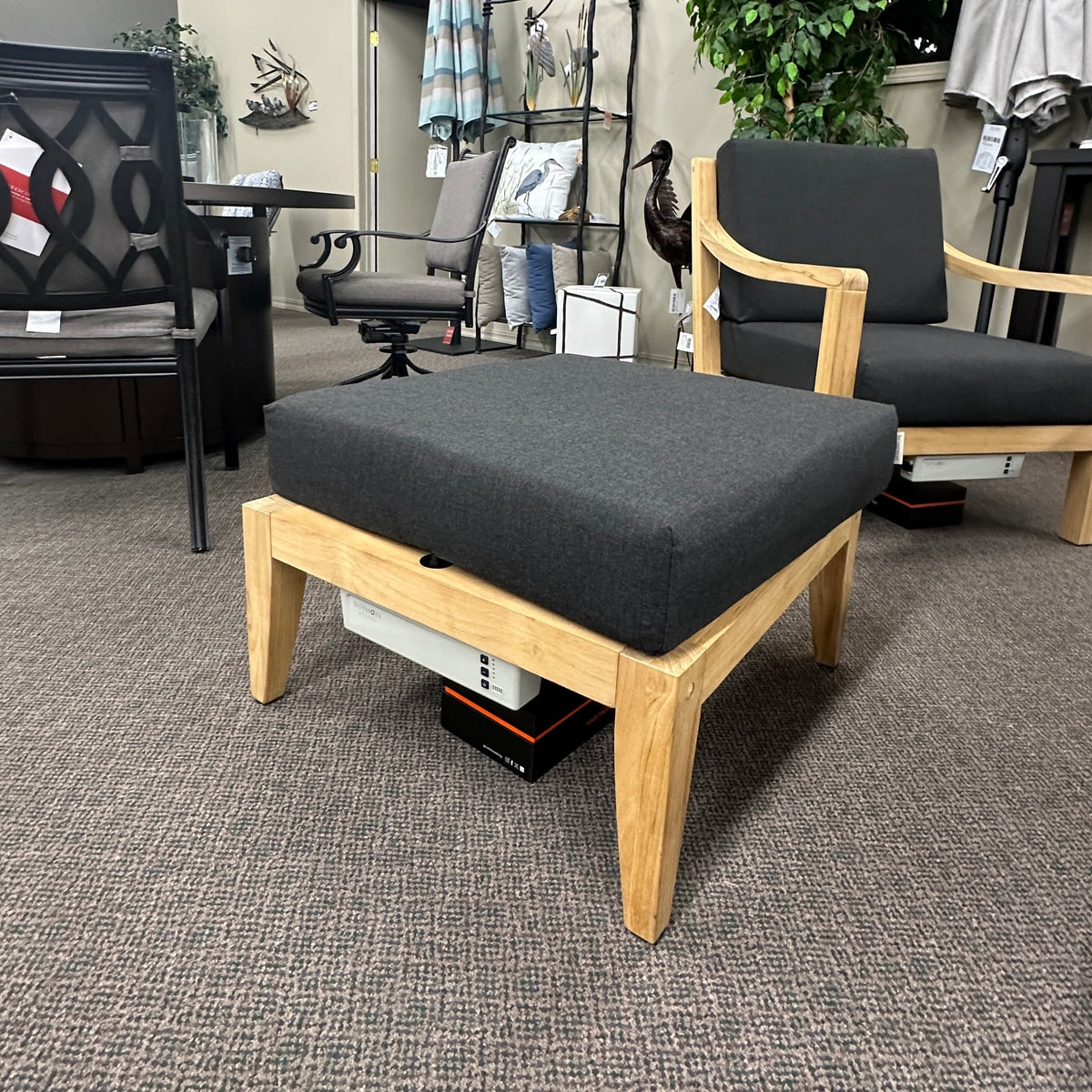 A wooden ottoman with a dark gray upholstered cushion is shown in a furniture showroom, positioned in front of two matching armchairs with light wood frames and dark gray cushions.