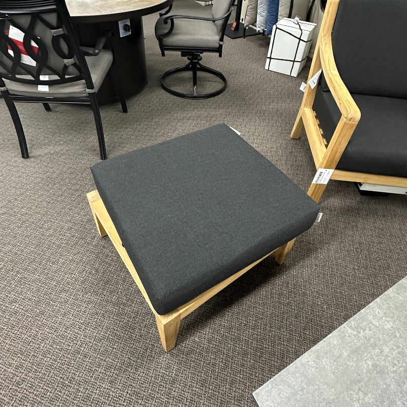 A modern wooden lounge ottoman with a dark gray upholstered cushion, shown in a furniture showroom with other seating pieces and decor.