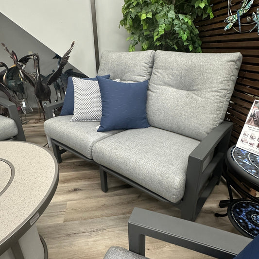 Outdoor patio furniture set with gray sofa, blue and white pillows, and a wooden wall with plants.