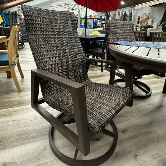 A dark brown wicker patio swivel chair with a high back and circular base, shown in a furniture store with a yellow patio umbrella and outdoor dining table in the background.