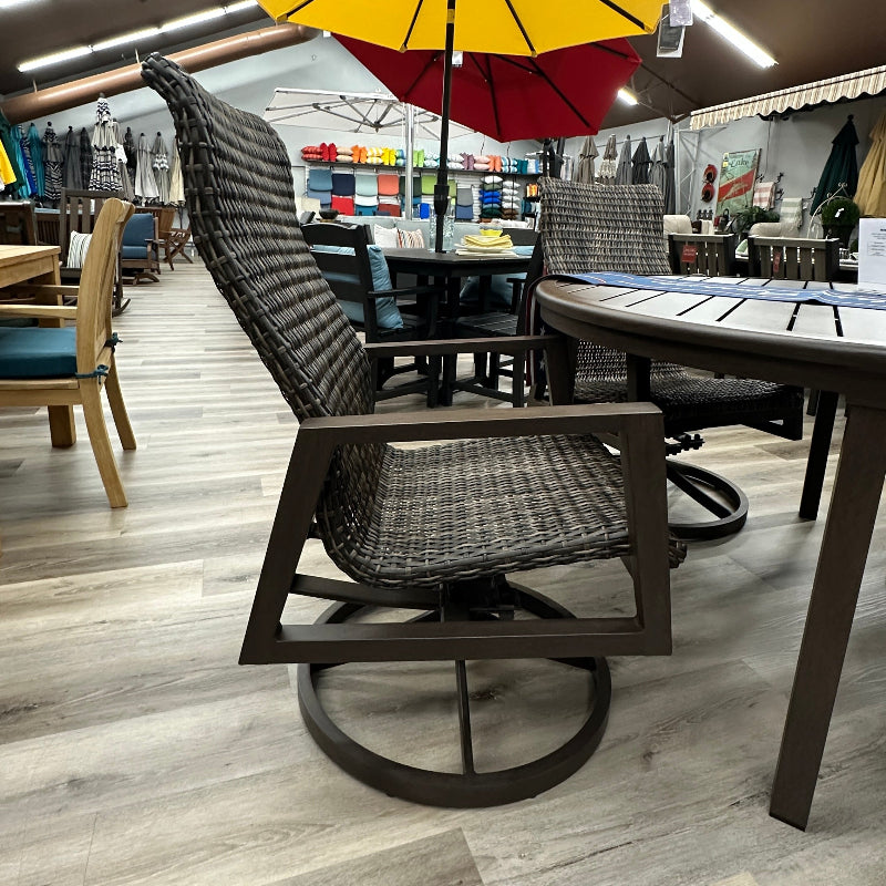 A dark brown outdoor swivel patio chair with a woven black and gray seat, positioned in a furniture store showroom with a yellow and red umbrella and a dining table in the background.