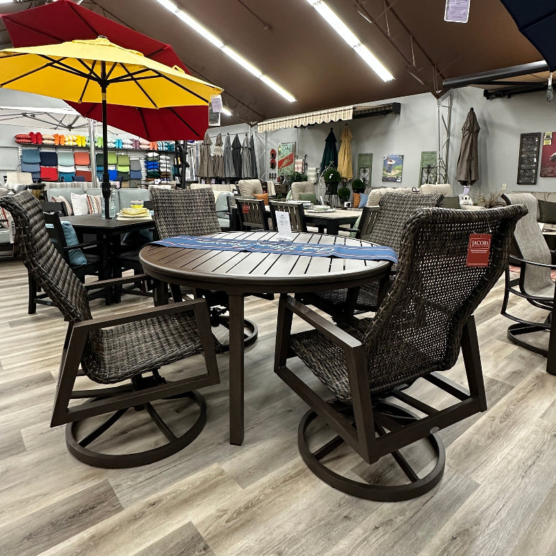 Outdoor dining set featuring a round dark brown table with a blue and white striped top, surrounded by four dark brown woven swivel chairs, in a furniture showroom with a red and yellow patio umbrella in the background.