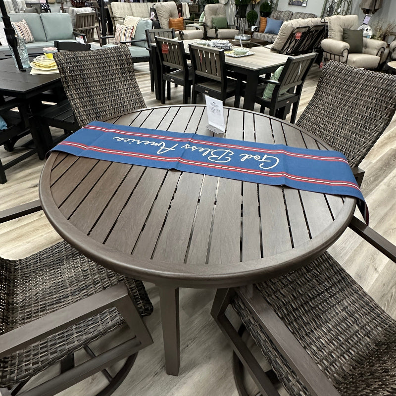 A round outdoor dining table set with four woven wicker chairs, featuring a blue and red runner across the table. The set is photographed inside a furniture showroom with other patio furniture and a patio umbrella in the background.