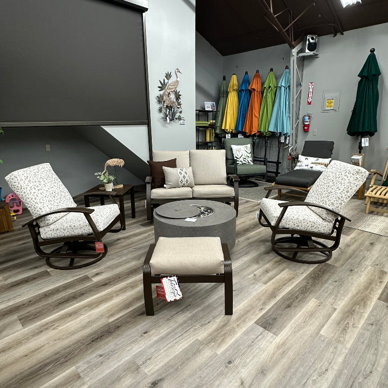 A showroom interior featuring outdoor patio furniture, including two patterned armchairs, a sofa, a round coffee table, and an ottoman, arranged on gray wood-look plank flooring. A large dark screen is mounted on the left wall, and colorful umbrellas are visible in the background.