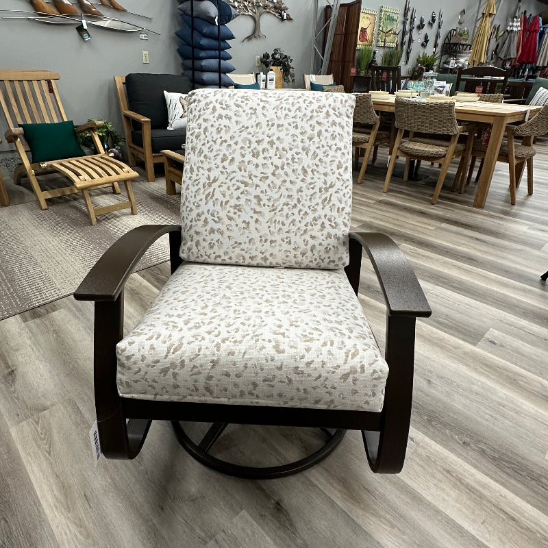 A modern swivel armchair with dark wood arms and base, upholstered in a light beige fabric with a small brown abstract floral pattern, shown in a furniture showroom.