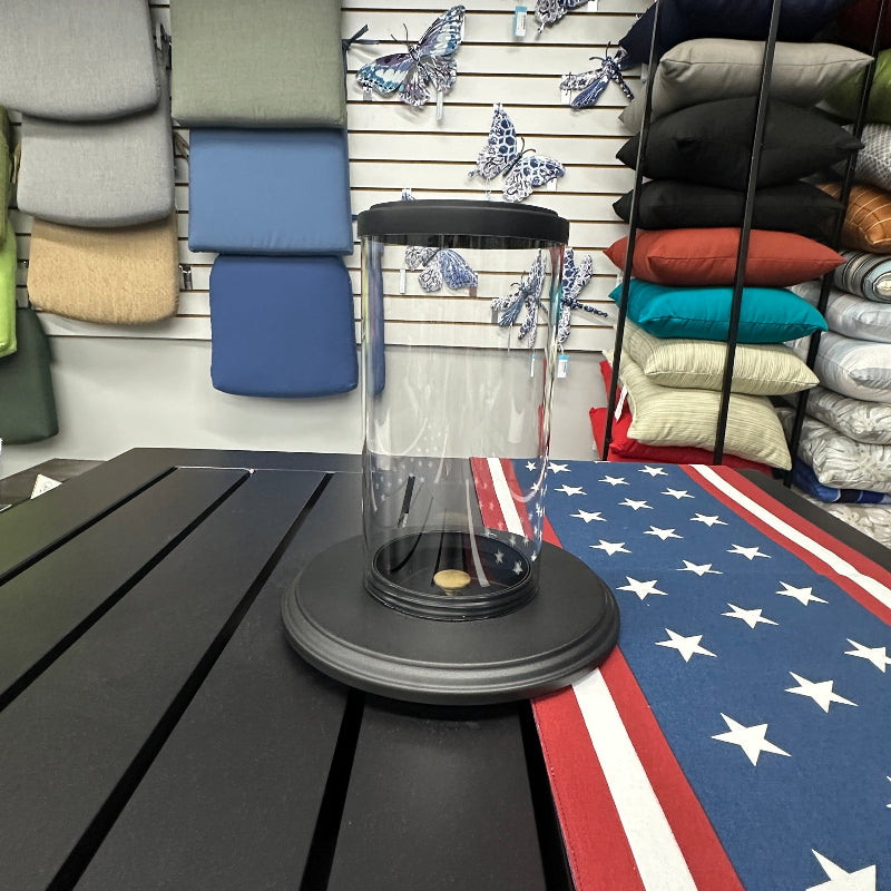 A clear cylindrical glass or acrylic lantern with a black top and base, displayed on a black slatted table in a retail showroom. The background shows folded textiles, fabric swatches, and an American flag-patterned mat on the floor.