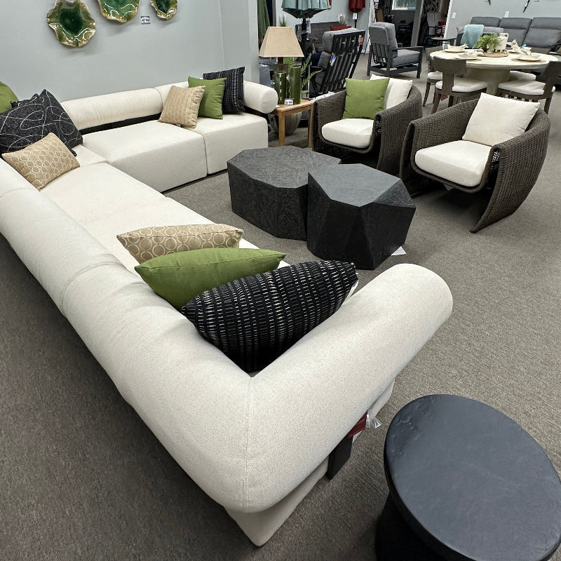 A modern outdoor sectional sofa in white with green and black patterned pillows, displayed in a furniture showroom alongside matching armchairs, coffee tables, and decor.
