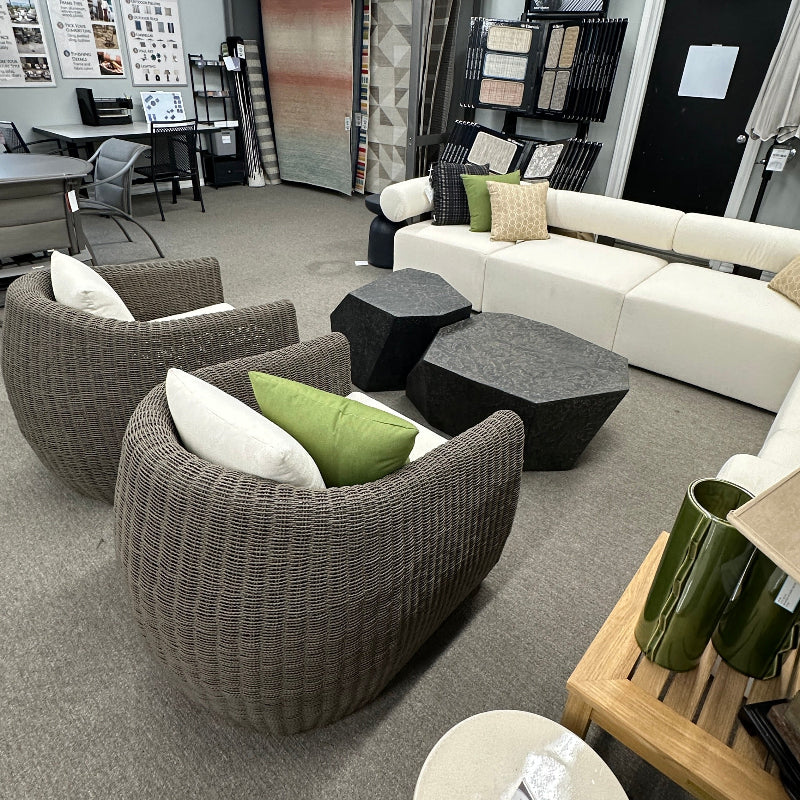 A showroom interior featuring two round woven armchairs with white and green pillows, a white sectional sofa, dark geometric coffee tables, shelving, and a carpeted floor.