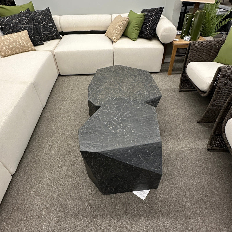 A modern living room setup featuring a white sectional sofa with green and beige accent pillows, a dark gray geometric coffee table in the foreground, and a gray carpeted floor.
