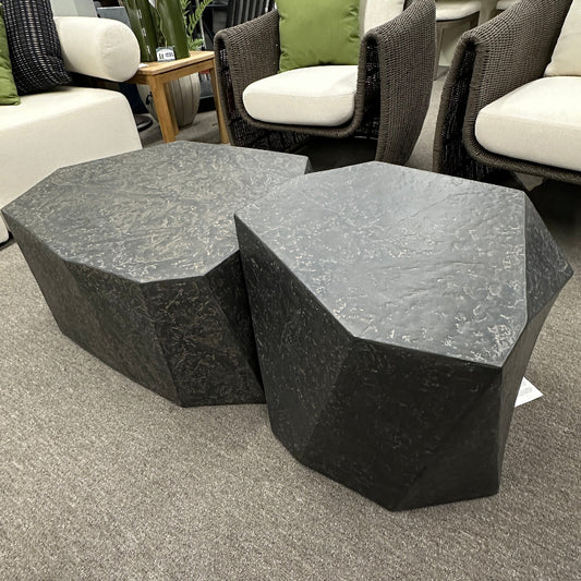 Two dark gray faceted stone side tables in a furniture showroom with gray carpet and outdoor chairs in the background.