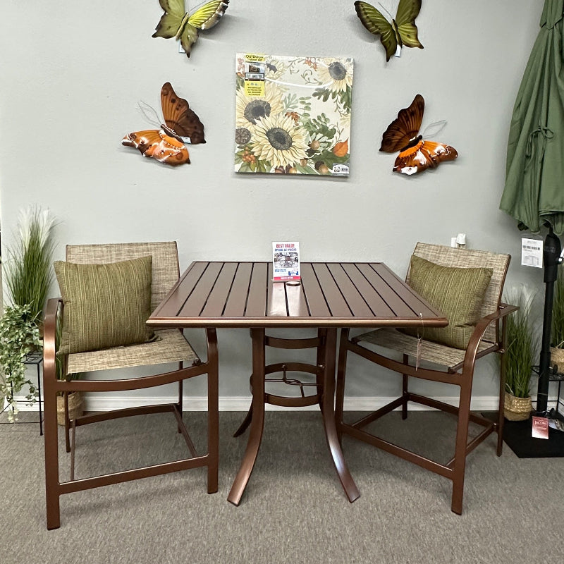 A 3-piece outdoor dining set consisting of a rectangular slatted table and two matching armchairs with olive green cushions, displayed in a showroom. The background wall is light gray with butterfly wall art and a sunflower print, and a closed green patio umbrella stands to the right.