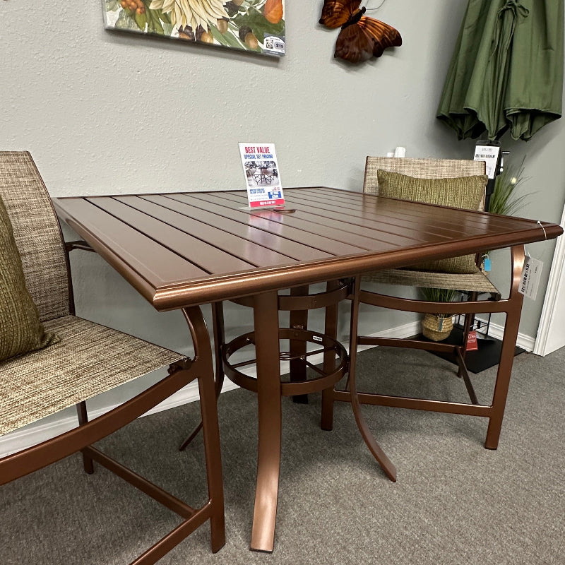 A rectangular outdoor patio dining table with a brown wood-look top and bronze-brown frame, shown in a showroom with matching chairs, a closed green patio umbrella, butterfly wall decor, and a floral print on the wall.